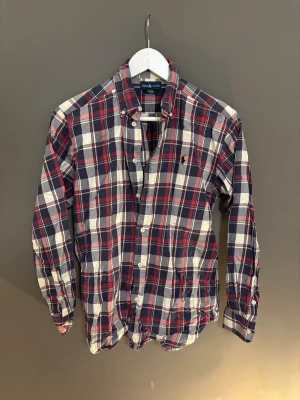 Rutig skjorta från Ralph Lauren  - Riktigt snygg rutig skjorta från Ralph Lauren i blått, rött och vitt. Skjortan har klassisk krage, knappar hela vägen och lång ärm. Tillverkad i 100% bomull för en skön känsla. Perfekt för dig som gillar preppy stil och vill sticka ut lite extra.