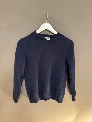 Mörkblå hoodie i kashmir från Soft Goat - Mysig mörkblå hoodie från Soft Goat i 100% kashmir. Tröjan har huva, långa ärmar och en klassisk, enkel design som passar perfekt till kyliga dagar. Supermjuk och skön att ha på sig, perfekt för dig som gillar stilrena plagg med lyxig känsla.