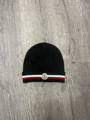 Svart Moncler mössa med ränder - Svart stickad mössa från Moncler med klassiska ränder i blått, vitt och rött längs kanten. Mössan har Moncler-logga framtill och är tillverkad i mjuk ull som håller dig varm. Perfekt accessoar för att lyfta din vinterstil.