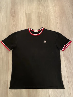 Svart Moncler t-shirt med röd/vit kant - Svart t-shirt från Moncler med korta ärmar och rund hals. Snygga röda och vita ränder på krage och ärmslut ger en sportig vibe. Moncler-logga broderad på bröstet. Tillverkad i mjuk bomull som känns skön mot huden.