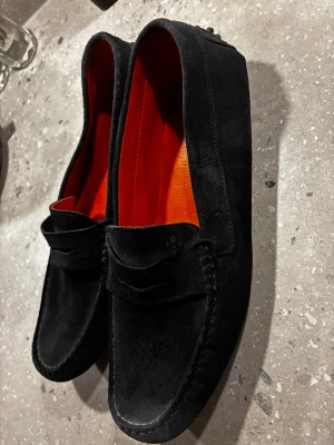 Morris - mörkblå mockaloafers med orange innersula - Säljer ett par stilrena loafers i mocka med klassisk pennyloafers-design. Skorna har en rund tå och en snygg orange innersula som ger en cool kontrast. Perfekta för dig som gillar en clean look med lite edge.