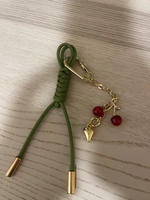 Vuitton Grön accessoar med körsbärsdetalj - Vuitton Snygg accessoar med grön flätad snodd och guldfärgade metalldetaljer. Utrustad med en karbinhake i guld och en kedja med röda körsbärsberlocker samt ett hjärta. Perfekt som nyckelring eller dekoration på väskan.