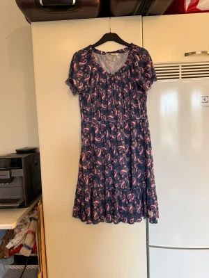 Blå paisleymönstrad klänning Odd Molly - Supersöt blå kortklänning från Odd Molly med paisleymönster i rosa och lila toner. Klänningen har korta ärmar, volangkant nertill och en avslappnad passform. Perfekt för dig som gillar boho-vibbar och vill ha något bekvämt men ändå snyggt. Från märket Odd Molly. Storlek 1 som enligt deras storleksguide motsvarar en S. Men passar mer en M. Rymlig. Mycket fint skick, jag hittar inga synliga anmärkningar eller defekter.  