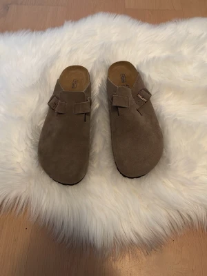 Birkenstock boston  - Säljer ett par bruna sandaler från Birkenstock i mjuk mocka med rund tå och justerbart spänne. Skorna har en platt sula med mönstrad undersida och bekväm fotbädd. Perfekt för dig som gillar chill och stilren look.
