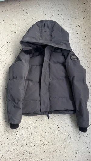 Mörkgrå pufferjacka från Canada Goose - Säljer en mörkgrå pufferjacka från Canada Goose med huva och snyggt broderat logomärke på ärmen. Jackan har dragkedja, knappar och ribbade muddar vid ärmslut. Insidan har meshfickor och praktiska axelband. Perfekt för kalla dagar!