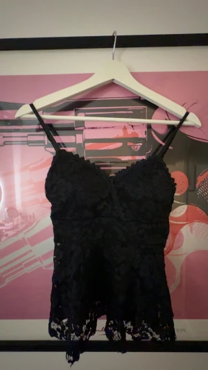 Svart spetsig linnetopp med smala band - Säljer en svart linnetopp med smala axelband och v-ringad hals. Toppen är täckt av spets med blommigt mönster och har en snygg kant nedtill. Perfekt att styla med jeans eller kjol för en cool look. Den har vadd i och är köpt på en marknad så vet ej storlek eller märke, den passar på mig som är Xs-s