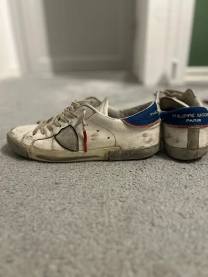 Philippe Model vita sneakers med blå detalj - Säljer ett par vita Philippe Model sneakers med blå häl och röda detaljer. Skorna har snörning, rund tå och platt sula. Tillverkade i skinn med beige och grå toner samt en diskret logga på tungan och hälen. Perfekt för dig som gillar fransk streetstyle.