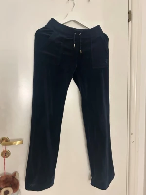 Svarta mjukisbyxor från Juicy Couture - Säljer ett par svarta mjukisbyxor från Juicy Couture med broderad logga på fickan och guldfärgade knappar bak. Byxorna har snörning i midjan, raka ben och är gjorda i mjuk sammet. Perfekta för chill dagar och har snygga fickdetaljer både fram och bak. DE ÄR UTSTRECHADE PÅ BILDERNA!!!