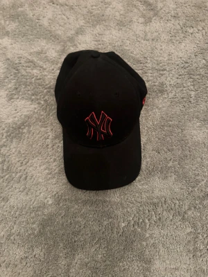 Svart New Era 9FORTY keps - Säljer en svart New Era 9FORTY keps med röd broderad NY-logga framtill och röd New Era-logga på sidan. Kepsen är justerbar baktill och tillverkad i 100% bomull. Perfekt för dig som gillar streetwear och sportig stil.
