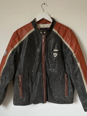 Mörkgrå och röd skinnjacka Tom Tailor - Cool mörkgrå skinnjacka från Tom Tailor med röda axlar och detaljer i beige. Jackan har ståkrage med tryckknappar, dragkedja framtill och två fickor med dragkedja. Märkespatchar på bröstet och ärmen ger en sportig vibe. Perfekt för dig som gillar retrostil. 7Y2