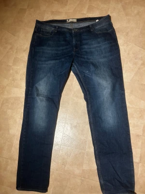 Mörkblå jeans med straight fit dressmann - Snygga mörkblå jeans med klassisk femficksdesign och raka ben. Jeansen har subtila slitningar och en straight passform som ger en avslappnad look. Tillverkade i robust denim med knapp och dragkedja framtill. Perfekt för dig som gillar enkel och tidlös stil.