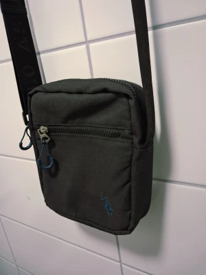 Svart axelväska från U.S. Polo Assn. - Säljer en stilren svart axelväska från U.S. Polo Assn. Väskan har justerbar rem med logotyp, ett huvudfack med dragkedja och en mindre ficka framtill. Materialet är slitstark polyester och väskan har en broderad logga samt en patch på framsidan.