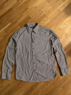 Randig grå skjorta från Tiger - Säljer en stilren grå skjorta med tunna vita ränder från H&M. Skjortan har klassisk krage, långa ärmar och knappar hela vägen framtill. Perfekt för dig som gillar enkel och clean stil. Materialet är mjuk bomull som känns skönt mot huden.