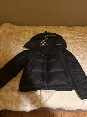 Svart pufferjacka med luva - Svart pufferjacka från Burberry med stor luva och klassiskt rutigt foder. Jackan har dragkedja och knappar framtill, två sidofickor med dragkedja och snörning vid huvan. Perfekt för kalla dagar och har en schysst, lite boxig passform.