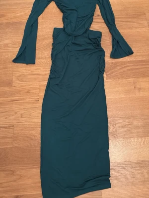 Mörkgrön långärmad långklänning - Snygg mörkgrön långklänning med långa ärmar och rynkade detaljer längs sidorna. Klänningen har en figurnära passform och är tillverkad i ett mjukt, stretchigt material som känns bekvämt mot huden. Perfekt för dig som vill ha en stilren och trendig look.