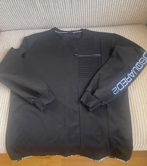 Svart sweatshirt från dsquard2 - Svart sweatshirt från Dsquard2 med ribbade muddar och coolt tryck på ärmen och nederkanten. Tröjan har en unik detalj med en ribbad panel och dragkedja på bröstet. Perfekt för dig som gillar streetwear och snygga loggor.