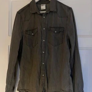 Grå denimskjorta från H&M - Snygg grå skjorta i denim från H&M med klassisk krage och två bröstfickor med knappar. Skjortan har långa ärmar och knäppning framtill med metallknappar. Perfekt för en chill och avslappnad stil.