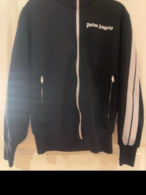 Palm Angels svart track jacket M - Svart track jacket från Palm Angels med vita ränder längs ärmarna och vit dragkedja. Tröjan har hög krage, två fickor med dragkedja och logga på bröstet. äkthets bevis på koftan finns!! 