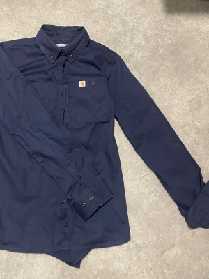 Mörkblå Carhartt skjorta med knappar - Snygg mörkblå skjorta från Carhartt med klassisk krage och knappar framtill. Bröstficka med Carhartt-logga och långa ärmar med manschettknappar. Tillverkad i en skön blandning av bomull och polyester med lite stretch för extra komfort. Den vita fläcken i hörnet tas bort innan fraktning. Vintage, därav lite dyrare men priset inte hugget i sten☺️