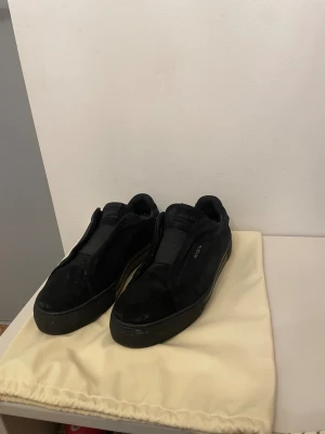 Svarta Axel Arigato sneakers i mocka - Säljer ett par svarta sneakers från Axel Arigato i storlek 45. Skorna är tillverkade i mjuk mocka med en platt sula och minimalistisk design. De har  elastisk panel istället för klassisk snörning. Tyvärr har lite av mockan på skorna skadats men det är inget man tänker på så mycket. Slå bara en pling 🛎️ kring några funderingar.