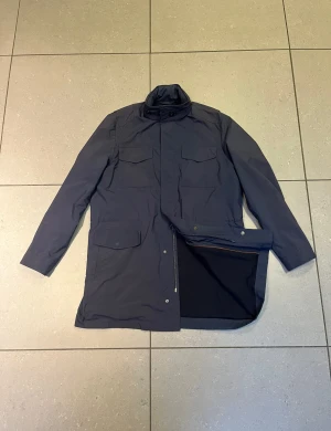 Mörkblå parkajacka från Loro Piana - Parkajacka i mörkblå färg från Loro Piana, tillverkad i Italien. Jackan har fyra stora fickor framtill, dragkedja och tryckknappar. Materialet är polyester på utsidan och mjuk ull på insidan. Perfekt för kyliga dagar med sin långa och stilrena design.