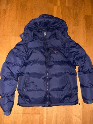 Mörkblå pufferjacka från Polo Ralph Lauren - Säljer en mörkblå pufferjacka från Polo Ralph Lauren med huva och dragkedja framtill. Jackan har en röd Polo-logga på bröstet, flera fickor med dragkedja och justerbara detaljer vid nederkant och ärmslut. Perfekt för kalla dagar och riktigt snygg stil. 