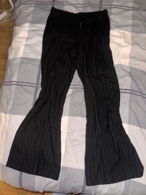 Svarta bootcut byxor från Brandy Melville - Säljer ett par svarta bootcut byxor från Brandy Melville med tunna vita ränder. Byxorna har dragkedja och knapp framtill, samt bälteshällor. Materialet känns som en mjuk bomullsblandning och passformen är loose med en snygg utsvängd siluett.