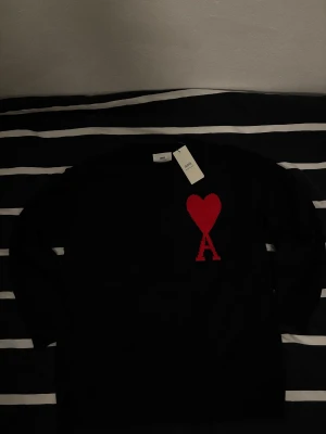 Svart stickad tröja från Ami - Svart stickad tröja från Ami Alexandre Mattiussi med ett stort rött hjärta och bokstaven A på bröstet. Rund hals med ribbad kant och långa ärmar. Perfekt för dig som gillar stilrena plagg med en twist. Rikigt skön nu på vintern, hör av dig om du har några frågor.