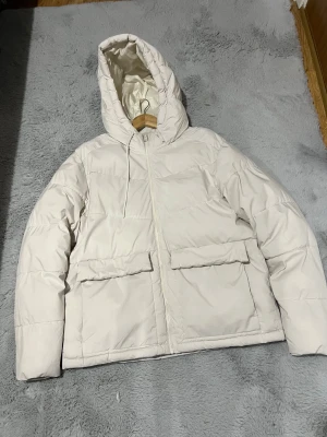 Vit pufferjacka från Jack & Jones - Säljer en clean och stilren vit pufferjacka från Jack & Jones i storlek L. Jackan har huva, två stora fickor framtill och dragkedja hela vägen. Materialet är polyester och jackan är perfekt för kalla dagar. Enkel design som funkar till många olika stilar.
