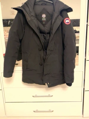 Svart dunjacka från Canada Goose M - Svart jacka från Canada Goose med klassisk logga på ärmen. Jackan har huva, dragkedja och knappar framtill. Perfekt för kalla dagar och har en stilren, längre passform. Materialet är slitstark polyester och dunfyllning som håller värmen.