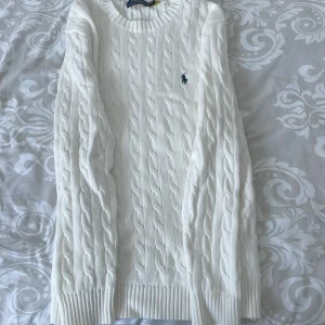 Vit kabelstickad tröja Polo Ralph Lauren - Vit kabelstickad tröja från Polo Ralph Lauren med klassisk rund halsringning och broderad marinblå logga på bröstet. Tröjan har ribbade muddar och är tillverkad i mjuk bomull. Perfekt för en clean och stilren look. (Helt ny!)