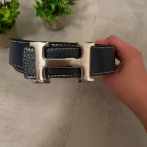 Blått bälte med H-spänne - Snyggt blått skärp med klassiskt stort H-spänne i silverfärgad metall. Bältet är gjort i äkta skinn med vit kontrastsöm längs kanterna och har en stilren, tidlös design som passar till många outfits.