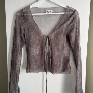 Sheer brun omlottopp från Weekday - Säljer en transparent grå/ grön topp från Weekday med långa ärmar och v-ringning. Toppen har knytning framtill och är croppad i modellen. Materialet är tunt och lätt, perfekt för lager-på-lager och en trendig look.