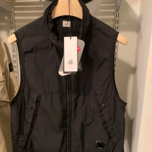 Svart dunväst från C.P. Company - Svart dunväst från C.P. Company med hög krage, dragkedja framtill och två snedställda fickor med knapp. Västen har den klassiska C.P. Company-linsen på ena fickan och är tillverkad i lätt syntetmaterial med Primaloft-foder. Perfekt för lager-på-lager och streetwear.