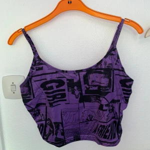 Lila croppad linne med tryck - Säljer ett lila croppat linne med svarta grafiska tryck och text. Linnet har tunna axelband och en tight passform, perfekt för att sticka ut. Materialet känns mjukt och stretchigt, vilket gör det bekvämt att bära hela dagen.