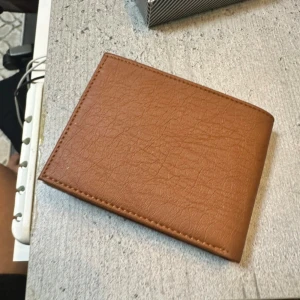 Brun plånbok från Leather Club - Snygg brun plånbok från Leather Club med klassisk design och flera fack för kort och sedlar. Tillverkad i äkta skinn med en naturlig struktur och stilrena sömmar. Perfekt storlek för att ha med sig överallt och har även ett transparent fack för ID eller foto.