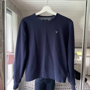 Mörkblå sweatshirt från GANT - Mörkblå sweatshirt från GANT med rund halsringning och broderad logga på bröstet. Tröjan har ribbade muddar vid ärmslut och nederkant och har en borstad insida. Barnstorlek 158/164 men sitter som en XS. 