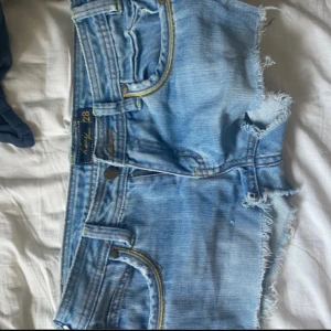 Ljusblå jeansshorts från Kenzie - Säljer ett par ljusblå jeansshorts från Kenzie med råa, fransiga kanter och klassisk femficksdesign. Shortsen har gul kontrastsöm och broderad logga baktill. Perfekta för varma dagar och har en avslappnad vibe.