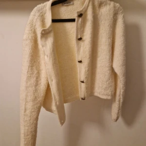 Krämvit kofta från Bisou med knappar - Mysig krämvit kofta från Bisou med fluffig textur och snygga knappar framtill. Koftan är långärmad och har en lite boxig, croppad passform. Perfekt att slänga på sig när det är kyligt ute.