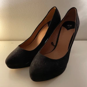 Svarta pumps med klack från H&M - Säljer ett par klassiska svarta pumps från H&M i storlek 39. Skorna har rund tå, hög blockklack och är klädda i mjuk mockaliknande material. Perfekta för dig som vill ha en stilren och tidlös look med lite extra höjd. Lite slitna på undersidan men påverkar inte skon särskilt mycket. 