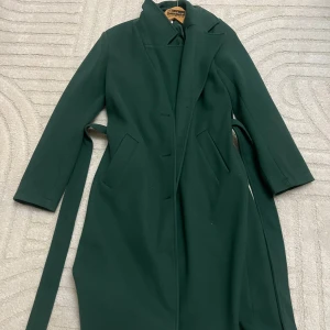 Mörkgrön kappa med bälte - Snygg mörkgrön kappa från Andiata med klassisk krage och bälte i midjan. Kappan har långa ärmar, dolda knappar framtill och två fickor. Perfekt för dig som vill ha en stilren och tidlös look under kyliga dagar.