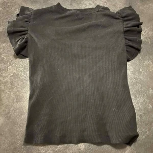 Svart ribbad topp med volangärmar - Svart ribbad topp med korta volangärmar som ger en snygg och trendig look. Toppen har en rund halsringning och är figurnära i modellen. 