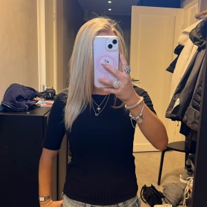 Svart ribbad topp med kort ärm - En svart, ribbad topp med rund hals och korta ärmar. Sitter figurnära och har en enkel, stilren look som passar till jeans eller kjol. Materialet känns mjukt och stretchigt, perfekt för en clean och snygg outfit.