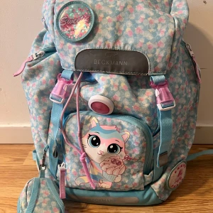 Pastellblå Beckmann ryggsäck med kattmotiv - Superfin pastellblå ryggsäck från Beckmann med blommigt mönster i rosa och vitt. Framsidan har en söt katt med cupcake och coola badges. Flera fack, vadderade axelremmar och reflexdetaljer. Regnskydd i neon medföljer. Perfekt för skolan eller utflykten!