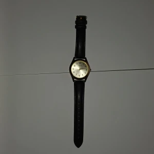 Guldig klocka från Regal med svart läderband - Stilren klocka från Regal med rund urtavla i guldton och tydliga svarta siffror. Klockan har ett klassiskt svart läderband och guldfärgat boett. Perfekt accessoar för dig som gillar enkel och tidlös design.