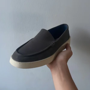 Mörkgrå loafers med vit sula - Snygga mörkgrå loafers med stilren design och kontrasterande vit platt sula. Skorna har rund tå och är tillverkade i slätt skinn, vilket ger en clean och modern look. Perfekta för dig som gillar minimalistisk stil.