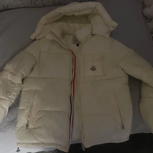 Vit Moncler pufferjacka med huva - Säljer en snygg vit pufferjacka från Moncler med avtagbar huva och klassisk logga på bröstfickan. Jackan har dragkedja med röd och blå detalj, två sidofickor med dragkedja och en bröstficka. Perfekt för kalla dagar och riktigt stilren. Pris kan diskuteras vid snabb affär.