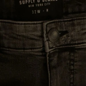 Grå slitna jeans Supply & Demand - Säljer ett par grå jeans från Supply & Demand med coola slitningar och färgstänk. Jeansen har normal passform, klassisk femficksdesign och knappstängning. Perfekta för dig som gillar en edgy streetstil.