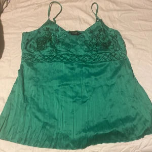 Grön glittrig satintopp Dorothy Perkins - Snygg grön satintopp från Dorothy Perkins med tunna axelband och v-ringning. Toppen har glittriga paljettdetaljer och broderier upptill samt ett satängliknande material som ger en lyxig känsla. Perfekt för dig som vill sticka ut med lite extra bling.