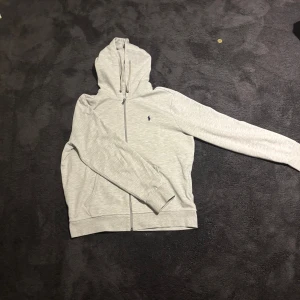 Grå hoodie från Polo Ralph Lauren - Snygg ljusgrå hoodie från Polo Ralph Lauren med dragkedja och klassisk logga broderad på bröstet. Tröjan har huva med snörning, långa ärmar och två fickor framtill. Perfekt för en chill och stilren look. Kan såklart gå ner i pris 